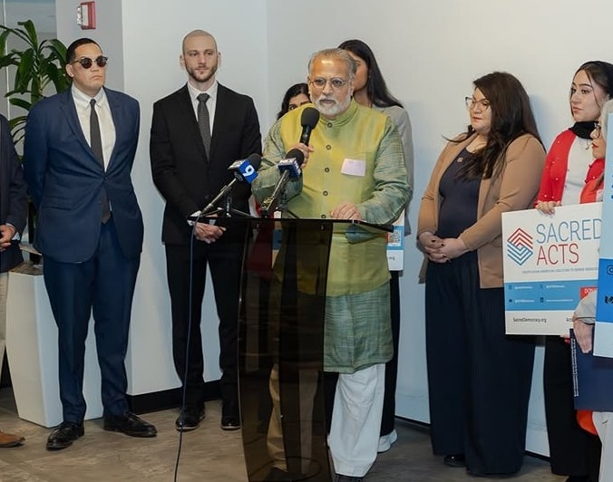 Tushar Gandhi at the podium at the 2026 Season for Nonviolence kickoff in Chicago, speaking into a bank of microphones with Sacred Acts and Sacred Democracy partners standing alongside him.