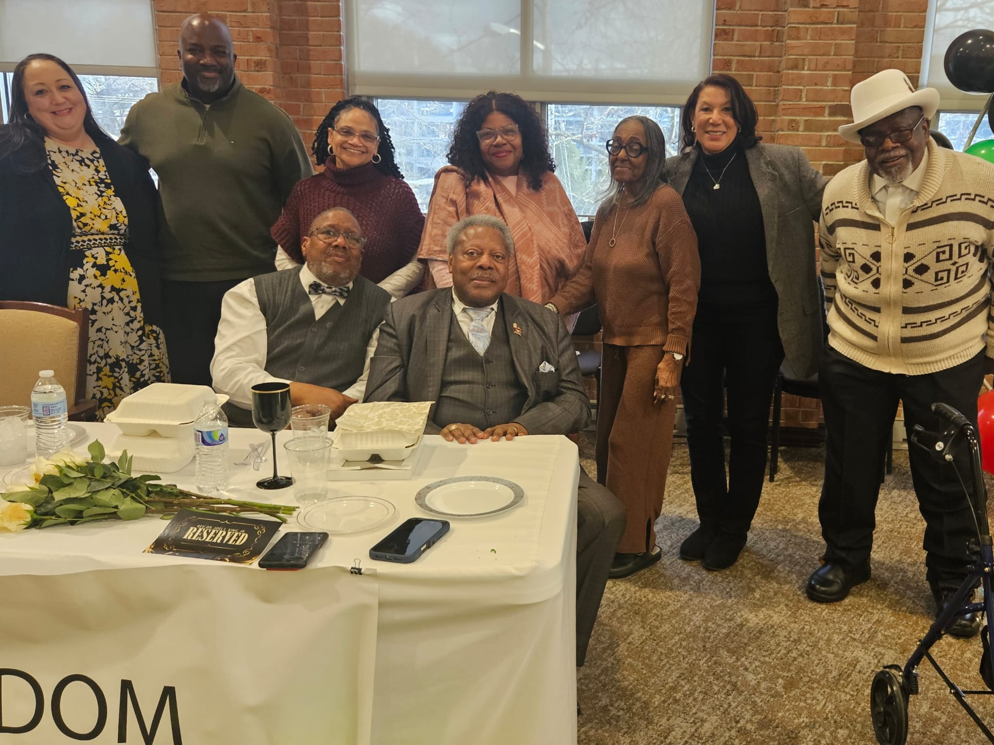 Rev. Joel King and Gregory Foster seated at a table with white roses, surrounded by family and community at a Black History Month gathering in Cincinnati, February 2026.