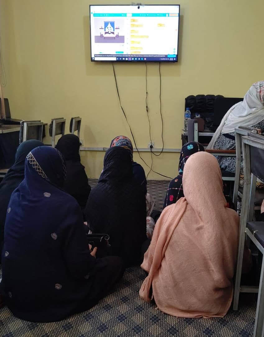A small group of students in headscarves sit on the floor, watching an mBlock visual-coding tutorial on a wall-mounted TV.